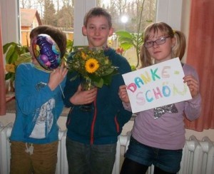 3 Kinder lächeln in die Kamera und halten Blumen, ein Luftballon und ein Dankeschön-Schild in der Hand