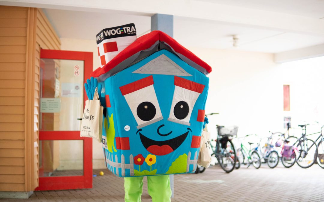 Ein Maskottchen in Form eines Hauses steht mit Jutebeteln vor einem Kindergarten.