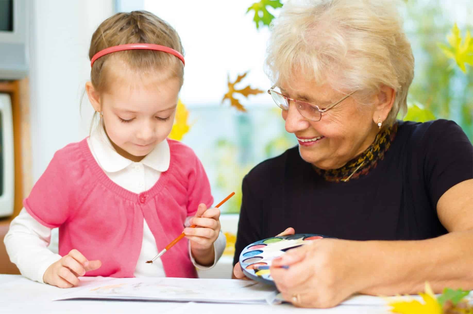 Eine ältere Frau malt gemeinsam mit einem jungen Mädchen am Tisch, Symbolbild für intergeneratives Lernen und Betreuung durch den Großelterndienst des SEFA e.V.