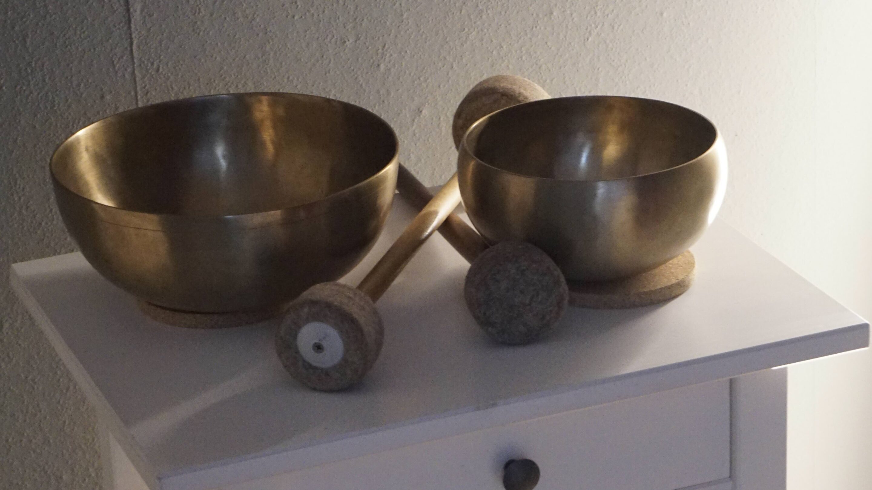 Zwei goldene Klangschalen und Klöppel auf einem weißen Tisch als Teil der ganzheitlichen Heilmethoden in der Praxis Dana Pfrötzschner.
